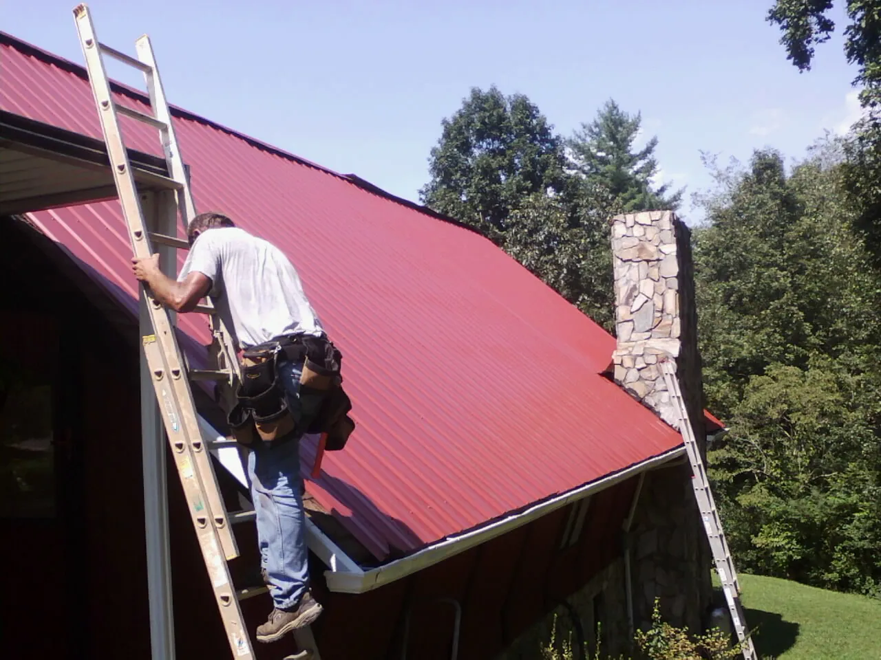 Roofer on a ladder beside a red metal roof and stone chimney — Soffit & Fascia Repair by Roofing Brothers in Mount Shasta